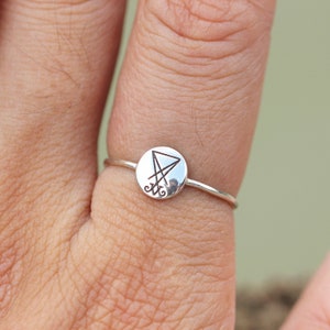 May include: A silver ring with a circular design featuring a sigil inside. The sigil is a triangle with a line through it and two curved lines at the bottom.
