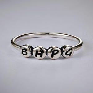 May include: A silver ring with four circular beads spelling out the letters "BHPG". The letters are in a bold, black font. The ring is on a white background.