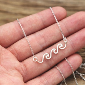 Puede incluir: Un collar de cadena de plata con un colgante de diseño de onda. El collar está en una mano.