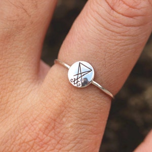 May include: A silver ring with a circular design featuring a pentagram symbol. The ring is worn on a finger.