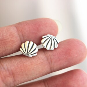 May include: A pair of small, silver-colored seashell stud earrings. Each earring features a detailed, fan-shaped design with radiating lines. The earrings are displayed on a person's hand.