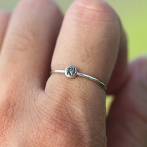 Puede incluir: Un anillo de plata con un pequeño dije redondo con la letra "R" grabada.
