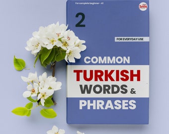 Puede incluir: Un libro azul con el título "Common Turkish Words & Phrases" y el número "2" en la portada. El libro está sobre un fondo morado claro con flores blancas.