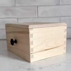 May include: A light-colored wooden box with a lid and a black knob. The box is constructed with dovetail joints and sits on a white surface. The background features a white brick wall.