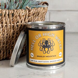Puede incluir: Un tarro de vela amarillo y negro con el texto "Hell or High Water" y una ilustración de un pulpo. La vela está etiquetada como "Bradley Mountain" y tiene una descripción de aroma de "Ron oscuro, madera flotante, vainilla ahumada".