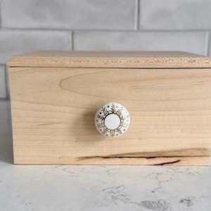 May include: A small, light-colored wooden box with a decorative knob. The knob is white with a gold and black snowflake design. The box has a simple, rectangular shape and a slightly curved top.