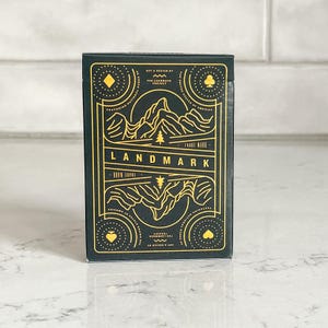 Könnte beinhalten: Ein Kartenspiel mit schwarzem Hintergrund und goldenen Akzenten. Die Schachtel zeigt eine Illustration einer Bergkette und den Text "LANDMARK - MADE MORE -  BORN EQUAL".