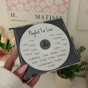 May include: A clear plastic CD jewel case containing a white disc with the text Playlist For Lexi and a list of twenty-six song titles printed in black ink. The disc features a central circular hole and a standard hub for secure placement.