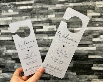 May include: Two white acrylic door hangers with a welcome message and a heart. The top hanger says "Welcome Sally & Sonny" and the bottom hanger says "Welcome Amanda & Shaun". Both hangers have a message that says "We're so excited to celebrate with you. Please enjoy your stay!"