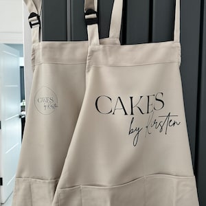 May include: Two beige aprons with adjustable straps and a large pocket. The apron on the right has the text "CAKES by Kirsten" printed on the front. The apron on the left has the text "CAKES by Kirsten" printed in a circle.