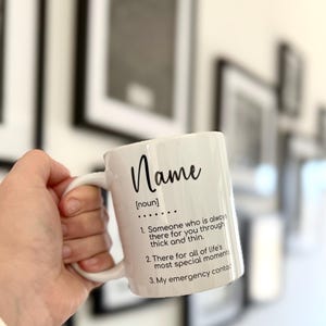 K&ouml;nnte beinhalten: Eine wei&szlig;e Keramiktasse mit einer schwarzen Aufschrift, die lautet: "Name [Substantiv] ... 1. Jemand, der immer f&uuml;r dich da ist, in guten wie in schlechten Zeiten. 2. Da f&uuml;r alle besonderen Momente des Lebens. 3. Mein Notfallkontakt:"