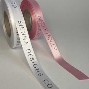 May include: Two rolls of satin ribbon, one white and one pink. The white ribbon has the text "SIENNA DESIGNS CO" printed on it. The pink ribbon has the text "NKD X HOLLY" printed on it.