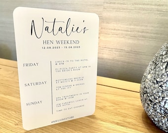 May include: A white card with black text outlining a weekend itinerary for a hen party. The card includes the name "Natalie's" at the top, followed by "Hen Weekend" and the dates "12.08.2023 - 15.08.2023". The card lists activities for each day of the weekend, including check-in to the hotel, a PJ pizza party, bottomless brunch, dinner and drinks, spa treatments, and a farewell lunch.