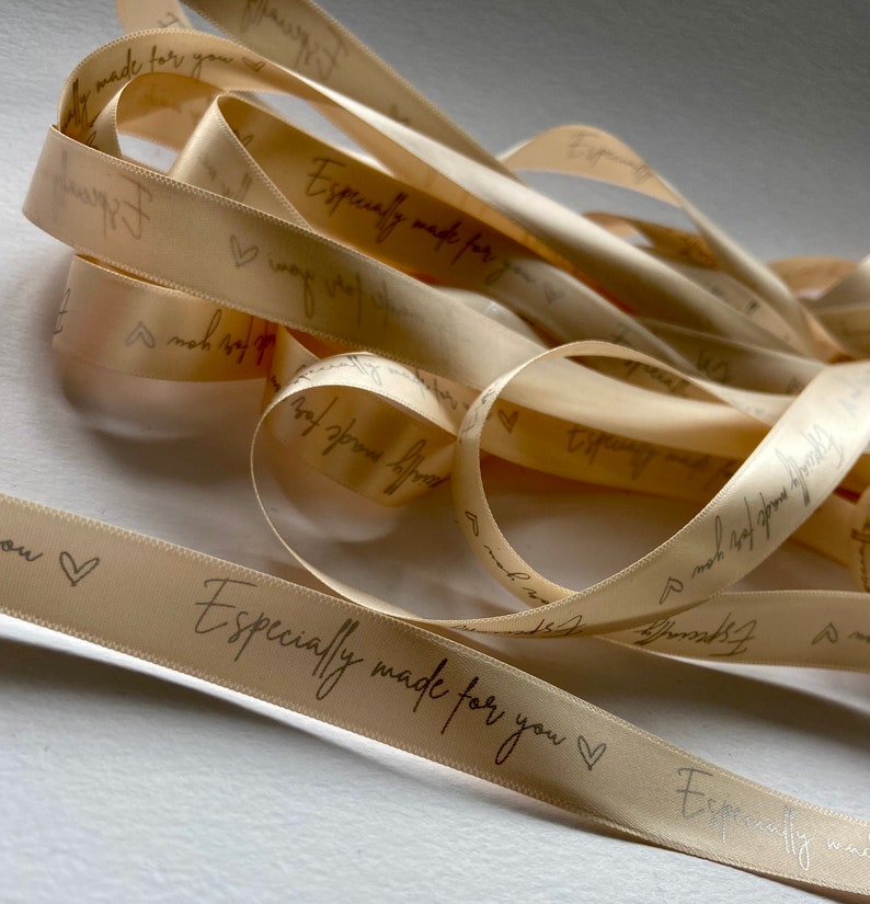 May include: A close-up of a spool of beige satin ribbon with the text "Especially made for you" printed in black ink. The ribbon is decorated with small hearts.