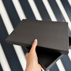 May include: A black square box with the lid slightly open, revealing the interior. The box is held against a background of alternating white and navy blue stripes.