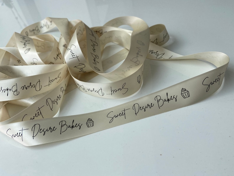 May include: A close-up of a white satin ribbon with the text "Sweet Desire Bakes" and a small cupcake icon printed on it.