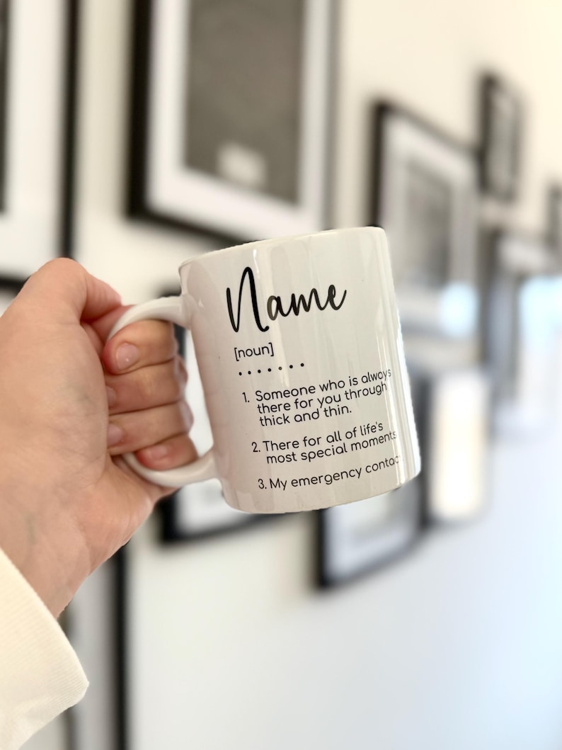 K&ouml;nnte beinhalten: Eine wei&szlig;e Keramiktasse mit einer schwarzen Aufschrift, die lautet: "Name [Substantiv] ... 1. Jemand, der immer f&uuml;r dich da ist, in guten wie in schlechten Zeiten. 2. Da f&uuml;r alle besonderen Momente des Lebens. 3. Mein Notfallkontakt:"