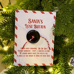 May include: A festive Christmas tree decoration. The card reads "Santa's Lost Button" and features a black button attached with red and white string. The card has red and white diagonal stripes and a poem about Santa's button.