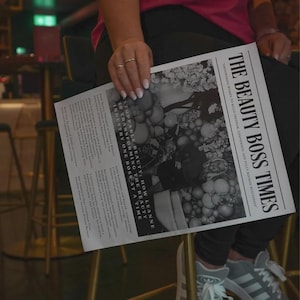 May include: A black and white newspaper titled "THE BEAUTY BOSS TIMES" is held by a person. The headline reads "EMPOWERING WOMEN TO BUILD FREEDOM, CONFIDENCE, AND BELLY BOOKED BUSINESSES". The paper features articles and images.