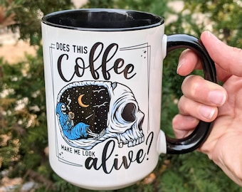 May include: White coffee mug with a black handle and rim. The mug has a black and white illustration of a skull with a skeleton inside drinking coffee. The text on the mug reads "Does this coffee make me look alive?"