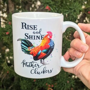 May include: White ceramic coffee mug with a colorful rooster illustration and the text "Rise and Shine Mother Cluckers".