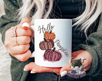 May include: White ceramic mug with a hand-painted design of three pumpkins in shades of orange, brown, and pink. The text "Hello Pumpkin" is written in a cursive font on the mug.