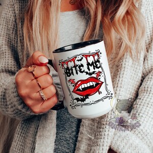 K&ouml;nnte beinhalten: Wei&szlig;e Keramiktasse mit schwarzem Rand und Henkel. Die Tasse zeigt eine schwarz-wei&szlig;e Illustration eines Vampirmundes mit Z&auml;hnen und dem Text "Bite Me" in einer schwarzen und wei&szlig;en Schrift.