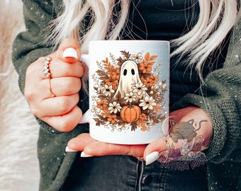 May include: White ceramic mug with a ghost and fall foliage design. The ghost is white and has a surprised expression. The foliage is in shades of orange, brown, and yellow. There are also small white flowers and a pumpkin.