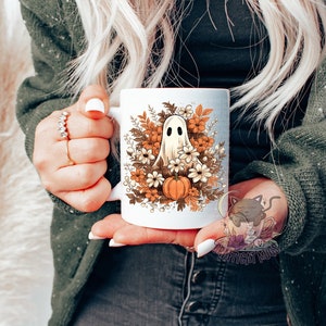 May include: White ceramic mug with a ghost and fall foliage design. The ghost is white and has a surprised expression. The foliage is in shades of orange, brown, and yellow. There are also small white flowers and a pumpkin.