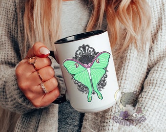 May include: A white ceramic mug with a black rim and handle. The mug features a green and pink luna moth with a black and white ornate frame. The mug has the text "Craftycat Mugs" on the bottom.