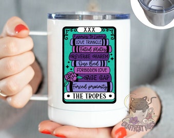 May include: White travel mug with a silver lid, featuring a graphic of stacked books with the text "Enemies to Lovers," "Love Triangle," and "The Tropes." The mug has a handle and is held by a person.
