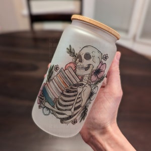 May include: A frosted glass jar with a bamboo lid features a hand-drawn illustration of a skeleton holding a stack of books. The skeleton is surrounded by flowers and leaves. The jar is a unique and whimsical way to store items.