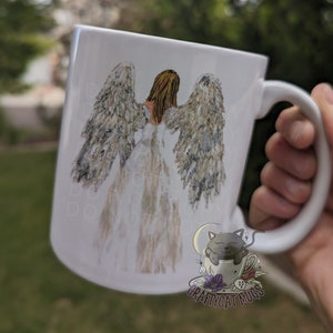 May include: White ceramic mug with a hand-painted design of an angel with wings. The angel is wearing a white dress and has long blonde hair. The mug also features a black cat sleeping in a teacup with a crescent moon above it. The text "CRAFTYCAT MUGS" is printed on the bottom of the mug.