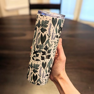 Puede incluir: Un vaso de acero inoxidable blanco con un patr&oacute;n floral con hojas verdes y flores moradas. El vaso tiene una tapa.