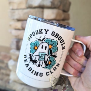 May include: White ceramic travel mug with a spooky ghost design. The ghost is holding a book that says "ghouls guide to witchery". The mug says "Spooky Ghouls Reading Club" in a black and white font.