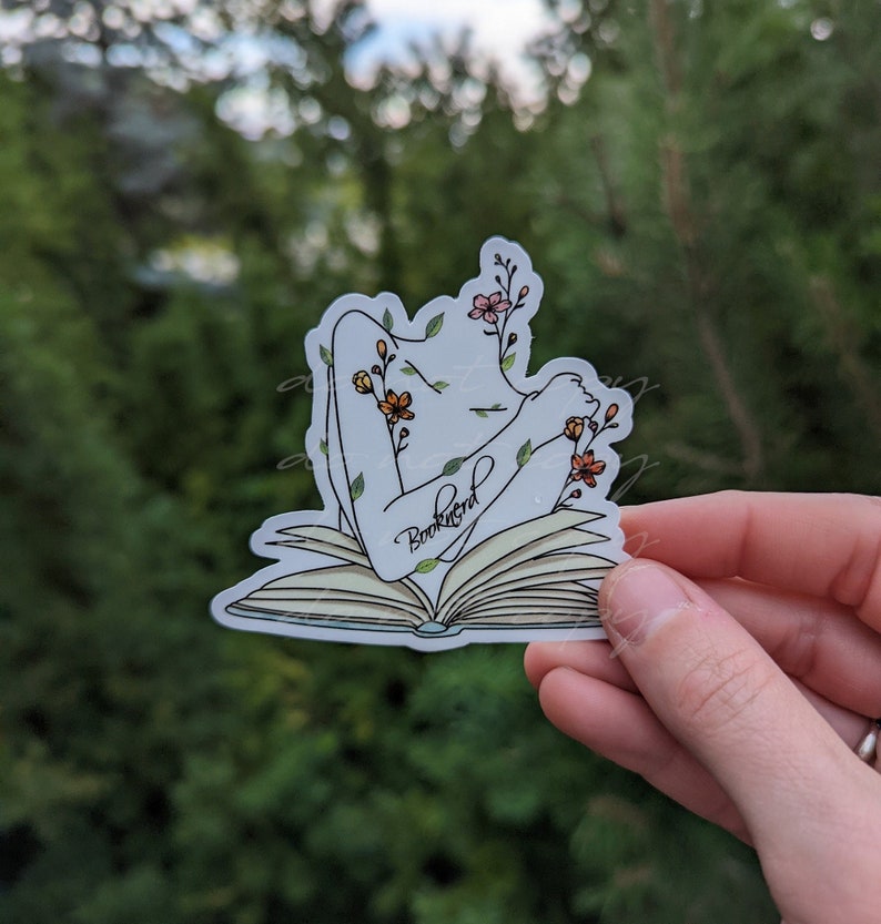 May include: A white sticker with a line drawing of a person with flowers growing from their body, sitting inside an open book. The book has the word "Booknerd" written on it.