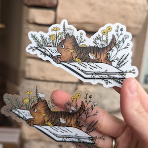 Op de afbeelding: Twee transparante stickers met een bruine tabby-kat die een boek leest, omringd door gele en witte bloemen. De kat ligt op het boek met zijn kop op de pagina's. De stickers zijn op een witte achtergrond.