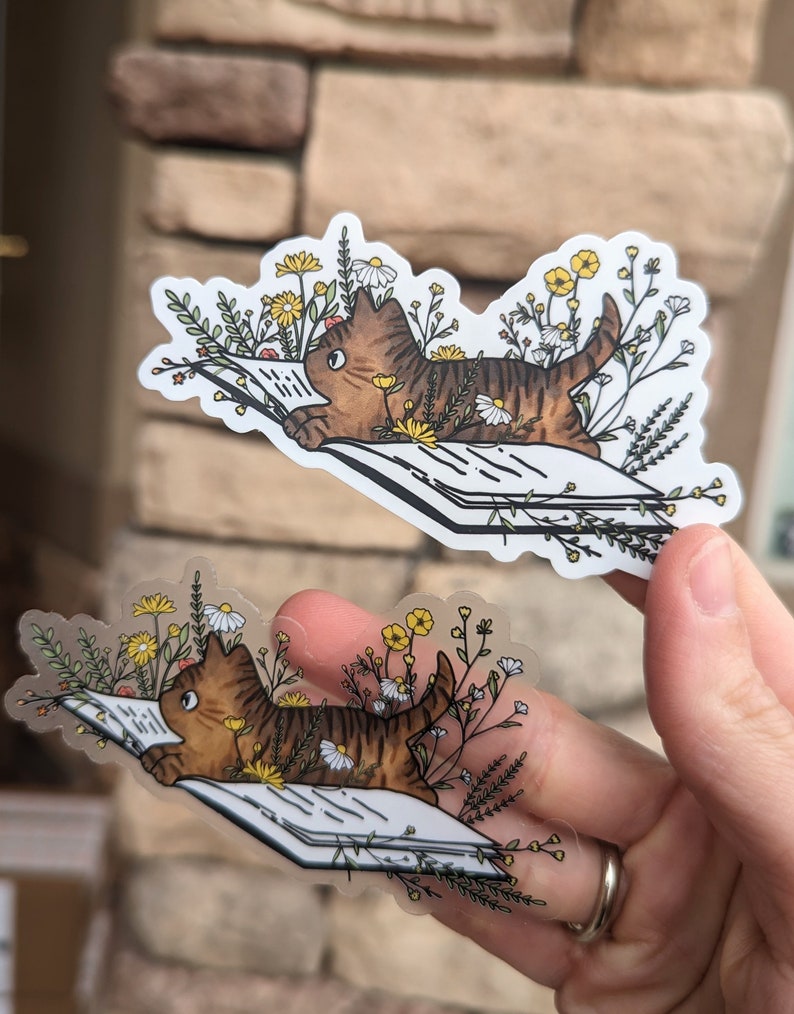 Op de afbeelding: Twee transparante stickers met een bruine tabby-kat die een boek leest, omringd door gele en witte bloemen. De kat ligt op het boek met zijn kop op de pagina's. De stickers zijn op een witte achtergrond.