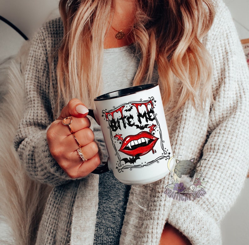 K&ouml;nnte beinhalten: Wei&szlig;e Keramiktasse mit schwarzem Rand und Henkel. Die Tasse zeigt eine schwarz-wei&szlig;e Illustration eines Vampirmundes mit Z&auml;hnen und dem Text "Bite Me" in einer schwarzen und wei&szlig;en Schrift.