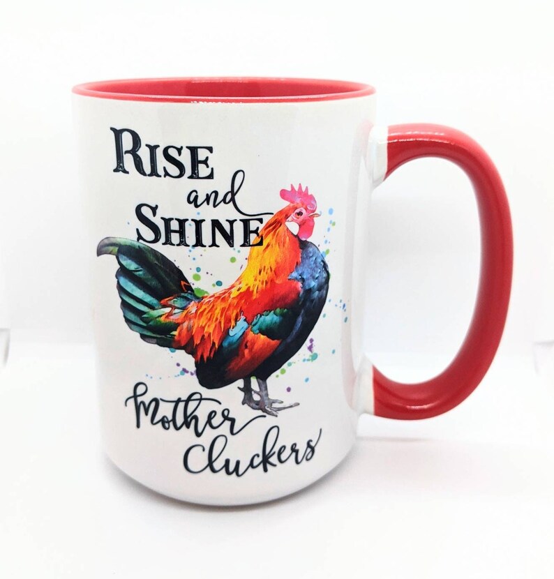 May include: White ceramic mug with a red rim and handle. The mug features a colorful rooster illustration and the text "Rise and Shine Mother Cluckers".