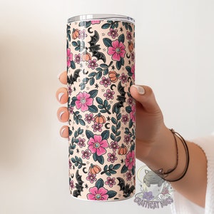 May include: A stainless steel tumbler with a cream background and a pattern of pink flowers, black bats, and orange pumpkins. The tumbler is 20 ounces and has a double-wall insulated design.