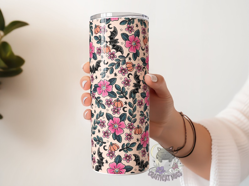 May include: A stainless steel tumbler with a cream background and a pattern of pink flowers, black bats, and orange pumpkins. The tumbler is 20 ounces and has a double-wall insulated design.
