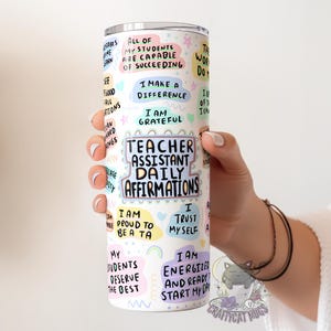 Op de afbeelding: Een witte roestvrijstalen tumbler met kleurrijke stickers met positieve affirmaties voor onderwijsassistenten. De tekst bevat "Teacher Assistant Daily Affirmations", "I am proud to be a TA", "I trust myself", en "I am energized and ready to start my day".