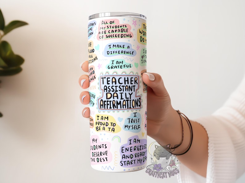 Op de afbeelding: Een witte roestvrijstalen tumbler met kleurrijke stickers met positieve affirmaties voor onderwijsassistenten. De tekst bevat "Teacher Assistant Daily Affirmations", "I am proud to be a TA", "I trust myself", en "I am energized and ready to start my day".