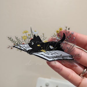Op de afbeelding: Een zwarte kat die een boek leest, omringd door gele en witte bloemen. Het boek is open en de kat ligt erop. De afbeelding is een transparante sticker.