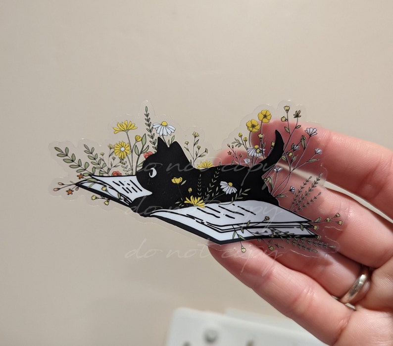 Op de afbeelding: Een zwarte kat die een boek leest, omringd door gele en witte bloemen. Het boek is open en de kat ligt erop. De afbeelding is een transparante sticker.