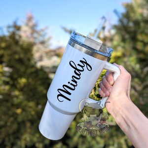 May include: White stainless steel travel mug with a black handle and a black inscription that reads "Mindy".
