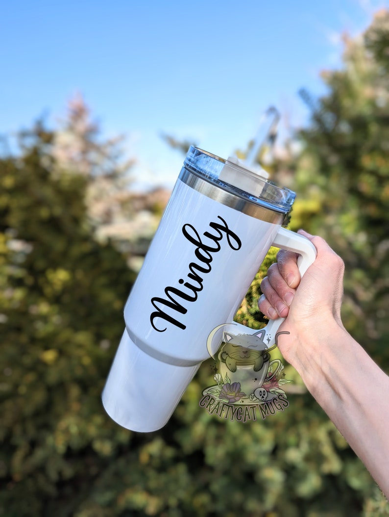 May include: White stainless steel travel mug with a black handle and a black inscription that reads "Mindy".