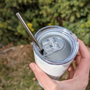 Puede incluir: Un vaso de acero inoxidable blanco con tapa transparente y una pajita de acero inoxidable. El vaso tiene una tapa transparente con un peque&ntilde;o anillo de silicona negro alrededor del borde.