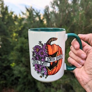 May include: White ceramic mug with a green rim and handle. The mug features a graphic of a pumpkin with a stitched-up face, half of which is a jack-o'-lantern. The pumpkin is surrounded by purple flowers and a banner that reads "Sorta Sweet" on one side and "Sorta Spooky" on the other.